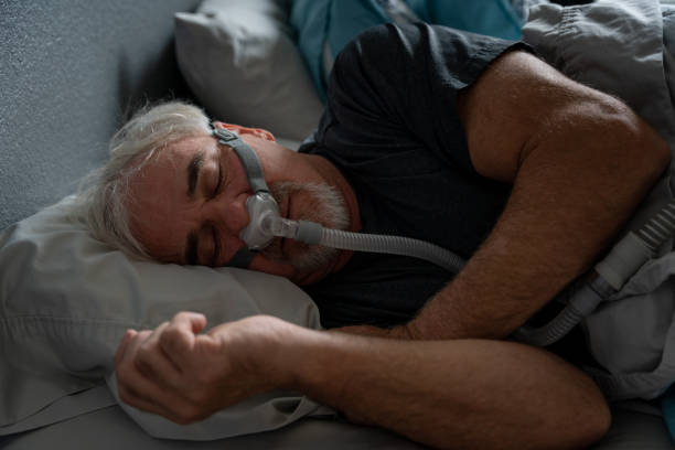 A man with gray hair and a beard sleeps on his side while wearing a CPAP mask.