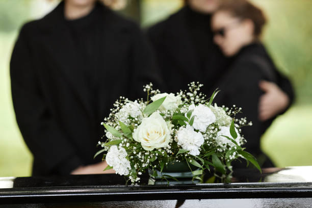 Close up of a white flower bouquet on coffin with people wearing black in the background.